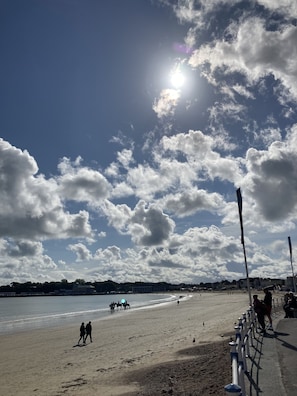 A beach with people walking and horseback riders in the distance, a clear sky, and a sun shining. - The Esplanade (Weymouth)