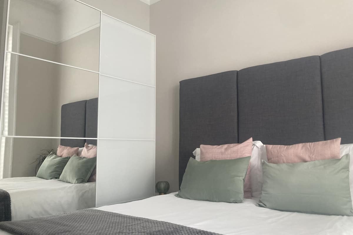 A bedroom with a large mirror, a grey headboard, and a bed with white bedding.