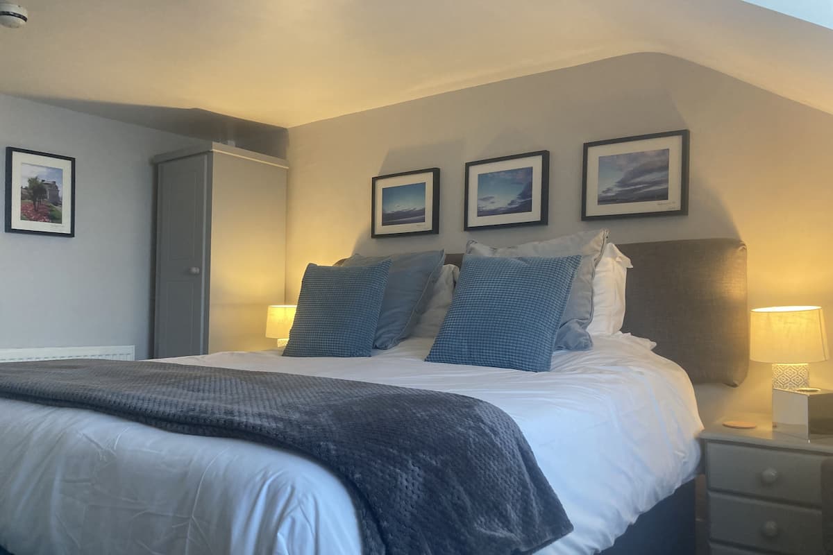 A neatly made bed with a white comforter and a dark gray blanket, flanked by two pillows. There are three framed pictures on the wall above the bed, and a nightstand with a lamp to the right.