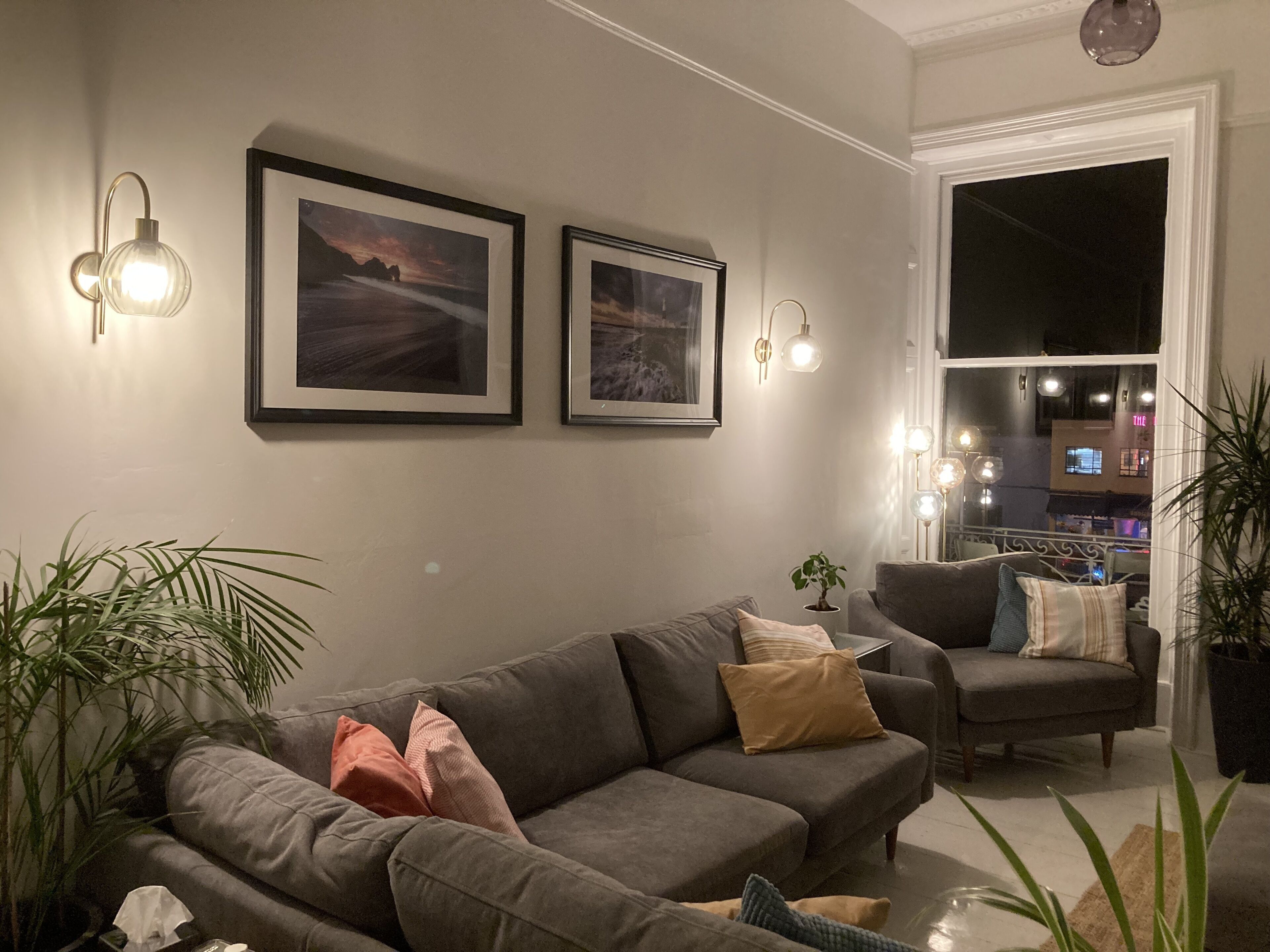 A living room with a grey sofa, a chair, and two framed pictures on the wall. There are potted plants and a window with a view of the city.