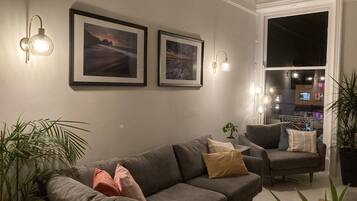 A living room with a grey sofa, a chair, and two framed pictures on the wall. There are potted plants and a window with a view of the city.