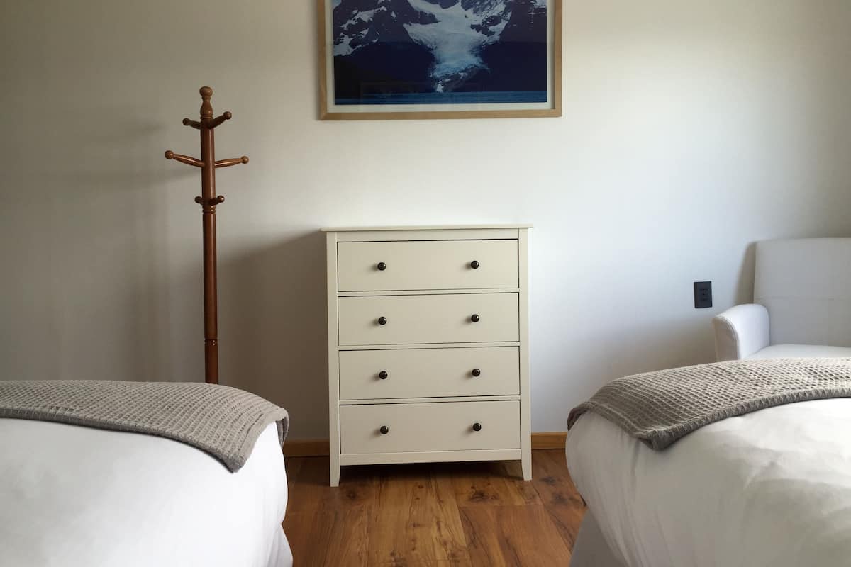 A bedroom with two beds, a wooden coat rack, a white dresser, and a framed picture of mountains.