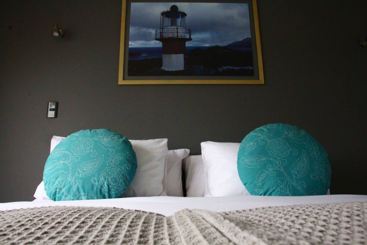 A bed with white bedding and teal patterned pillows, a framed picture of a lighthouse on the wall, and a textured blanket on the bed.