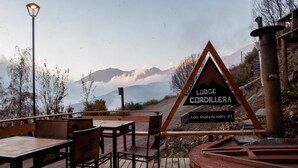 A wooden deck with tables and chairs, a sign for 'Lodge Cordillera Los Guayacanes 81', and a mountainous backdrop. - Lodge Cordillera (Lo Barnechea)