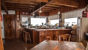 A rustic kitchen with wooden beams, a large island with stools, and a view of the outside. - Lodge Cordillera (Lo Barnechea)