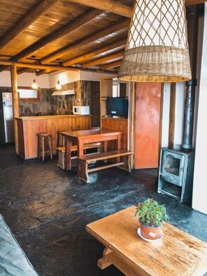 A rustic interior with a wooden table, a bench, a hanging light fixture, a stone floor, and a wood-burning stove. - Lodge Cordillera (Lo Barnechea)