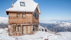 A wooden chalet with a snow-covered roof, large windows, and a view of snow-covered mountains. - Lodge Cordillera (Lo Barnechea)