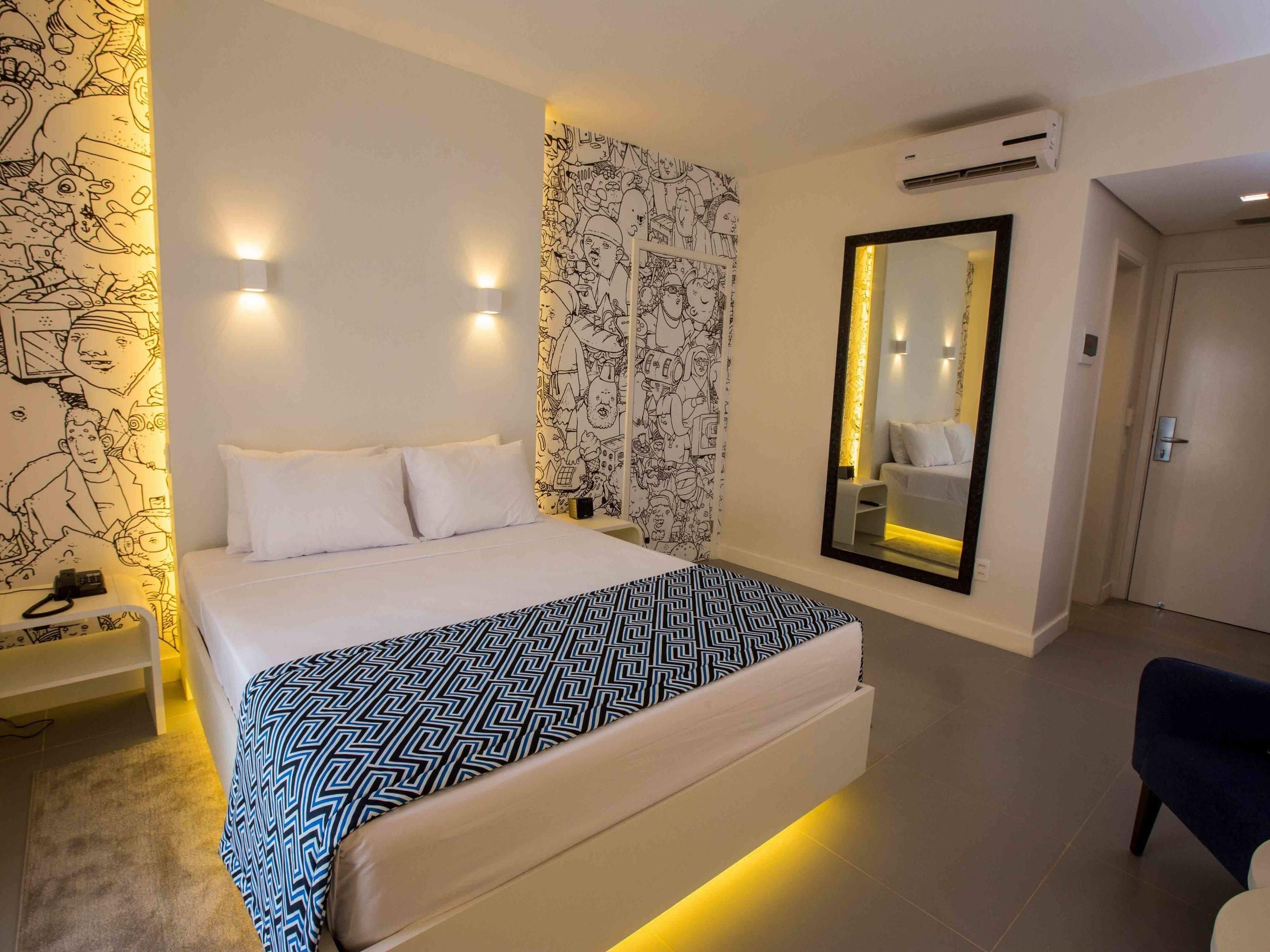 Ett modernt hotellrum med en stor säng, ett nattduksbord med en telefon, en mönstrad pläd, en vägg prydd med en svartvit väggmålning och en stor spegel.