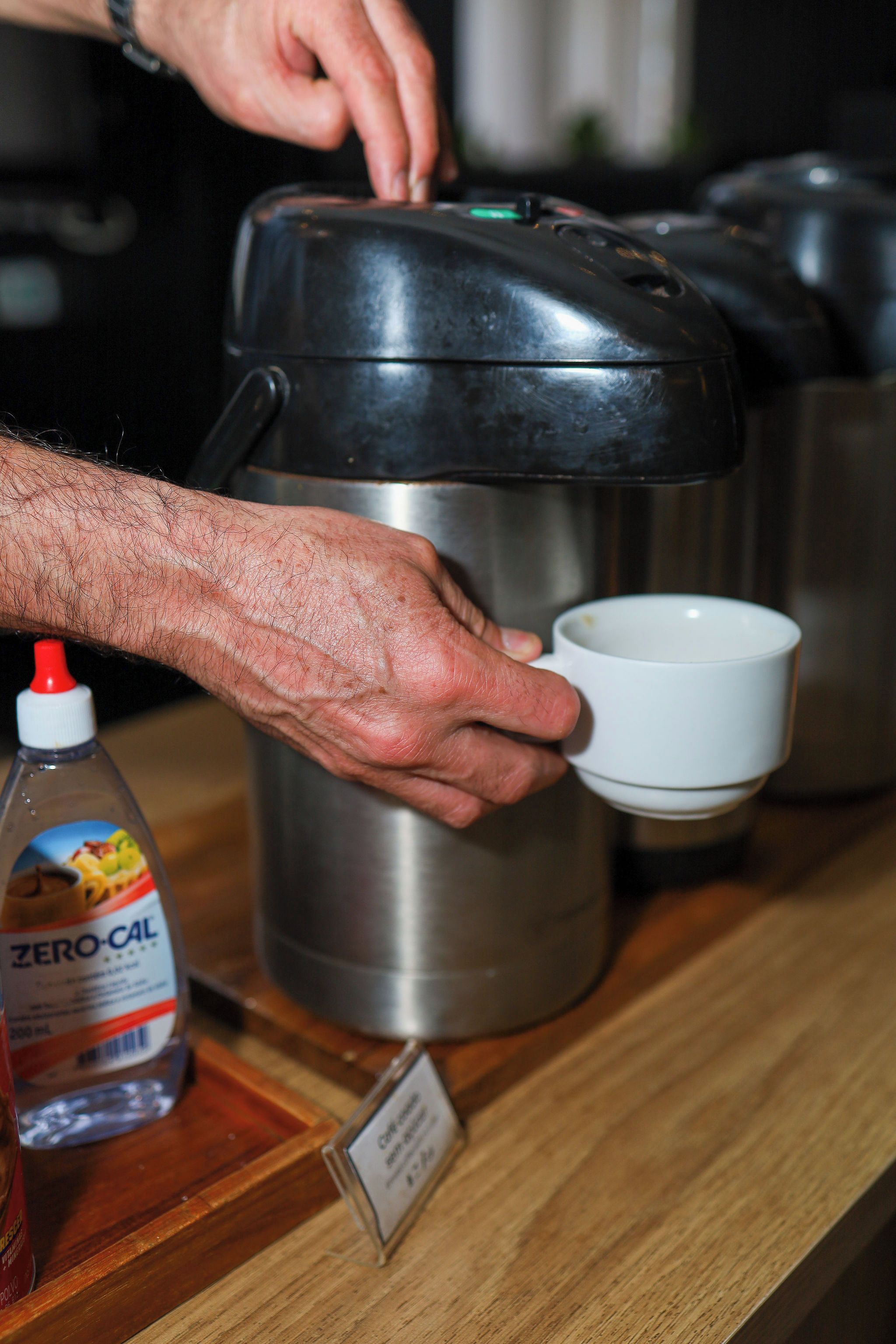 Una mano sosteniendo una taza blanca al lado de una tetera eléctrica de acero inoxidable sobre una encimera de madera.