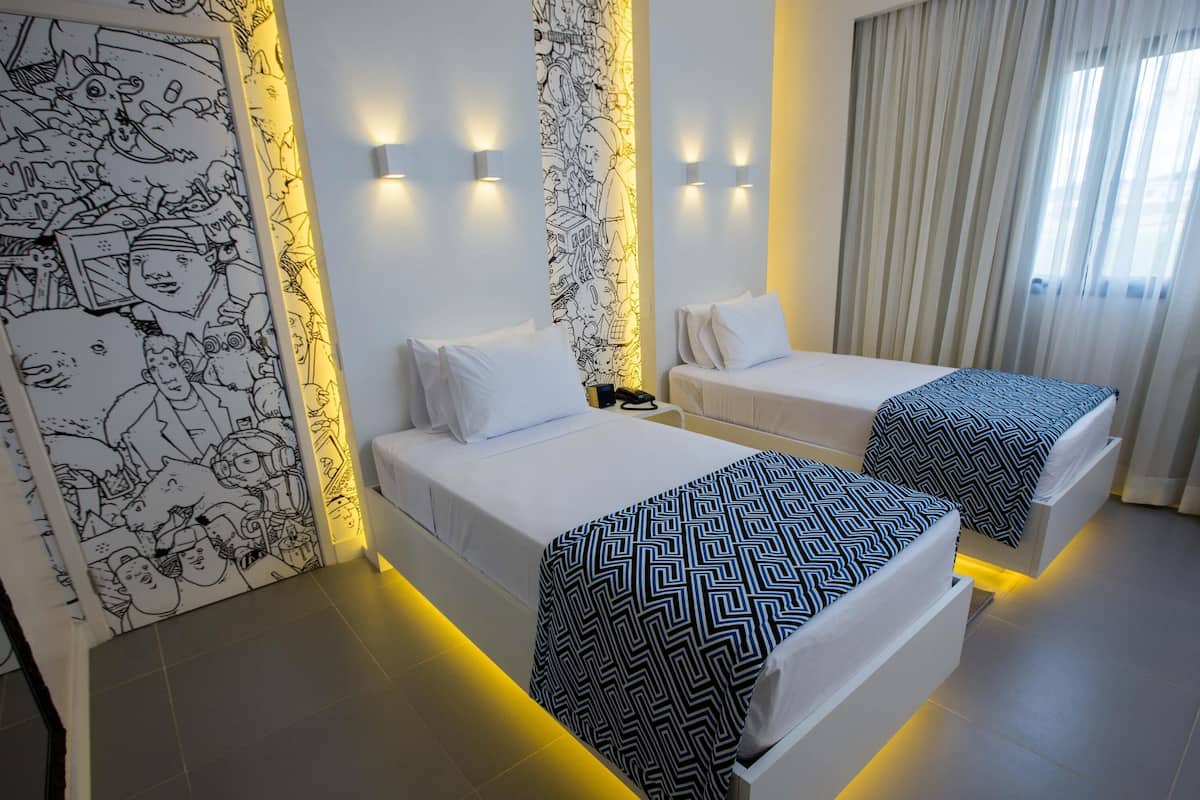 Une chambre d’hôtel avec deux lits, un mur orné d’une fresque en noir et blanc, et une fenêtre avec des rideaux.