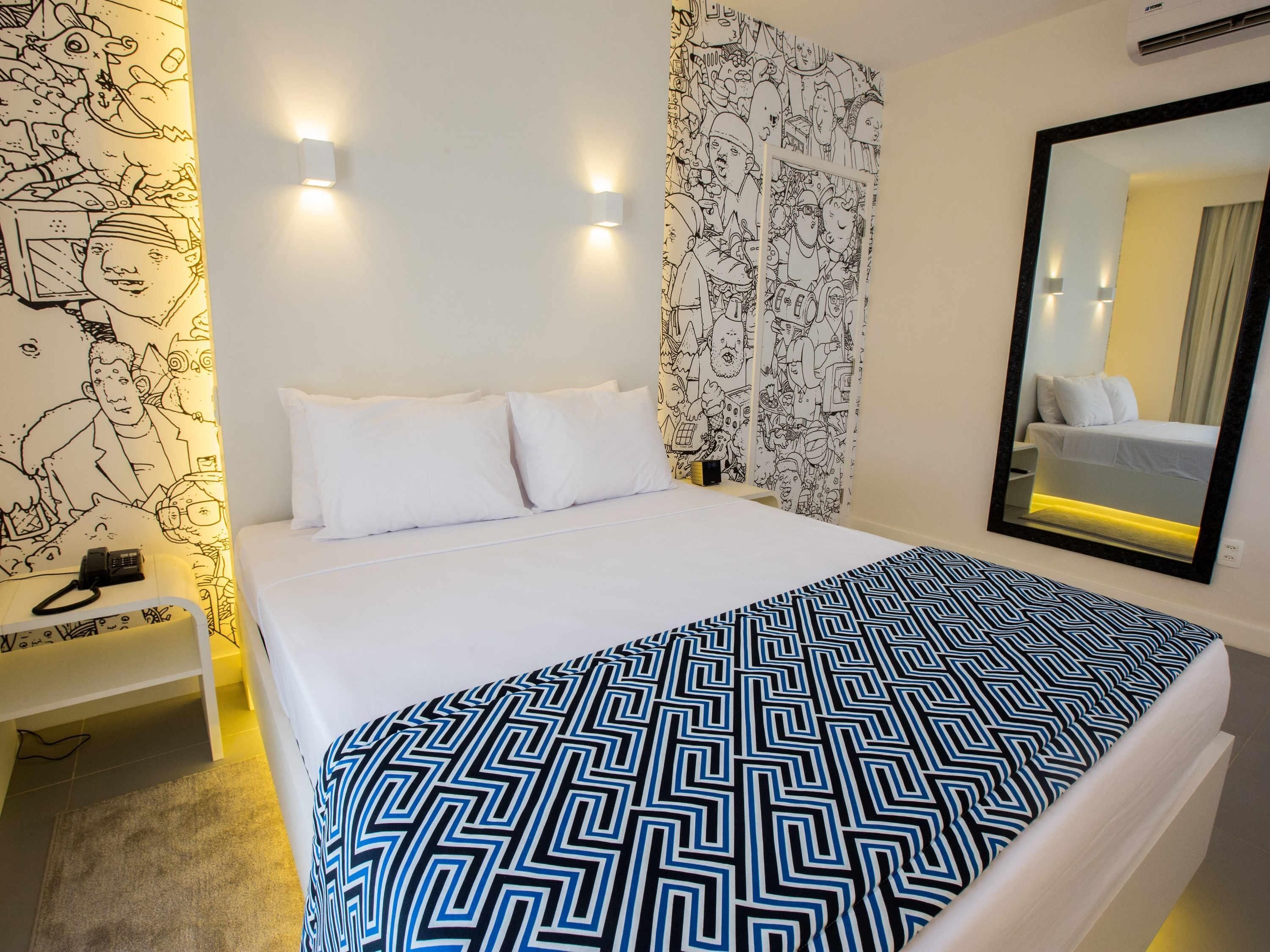 Habitación de hotel con cama, almohadas blancas, manta estampada, mesita de noche con teléfono y pared con espejo y obra de arte.