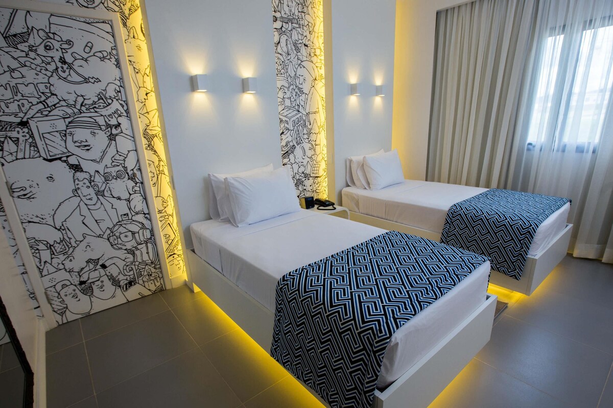Quarto de hotel com duas camas, uma parede com mural em preto e branco, e uma janela com cortinas.