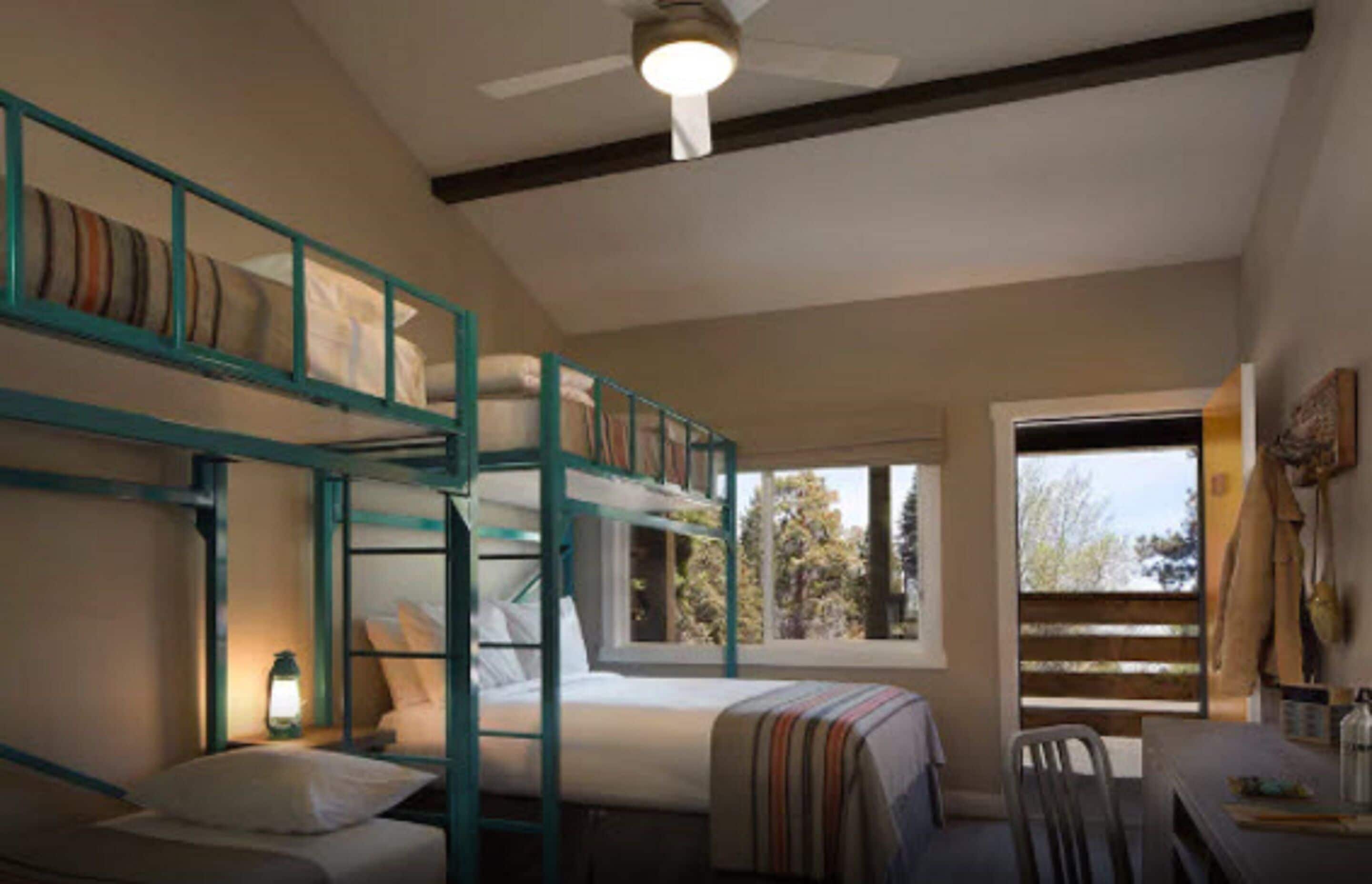 A bunk bed room with a ceiling fan, a window with a view of trees, and a desk with a chair.