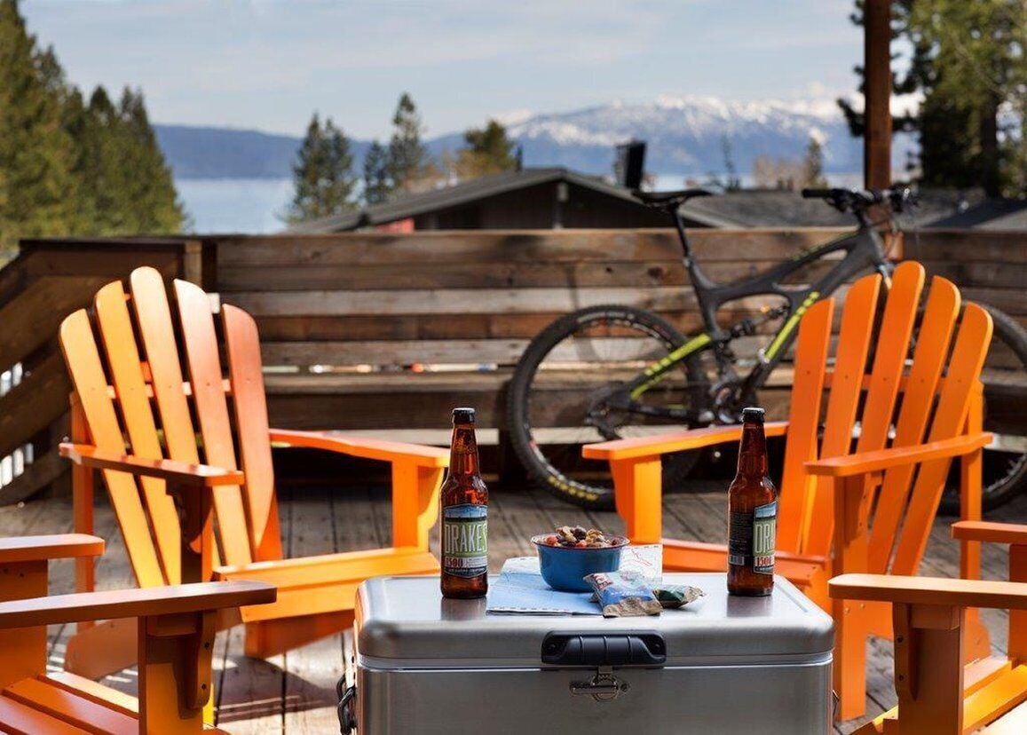 Two orange Adirondack chairs on a wooden deck with a bicycle and a suitcase in the background.