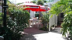 Een terras met een tafel en stoelen onder een rode parasol, omgeven door groen en een witte hek.