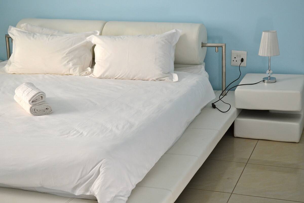 A neatly made bed with white linens and pillows, a white bedside table with a lamp, and a power outlet.