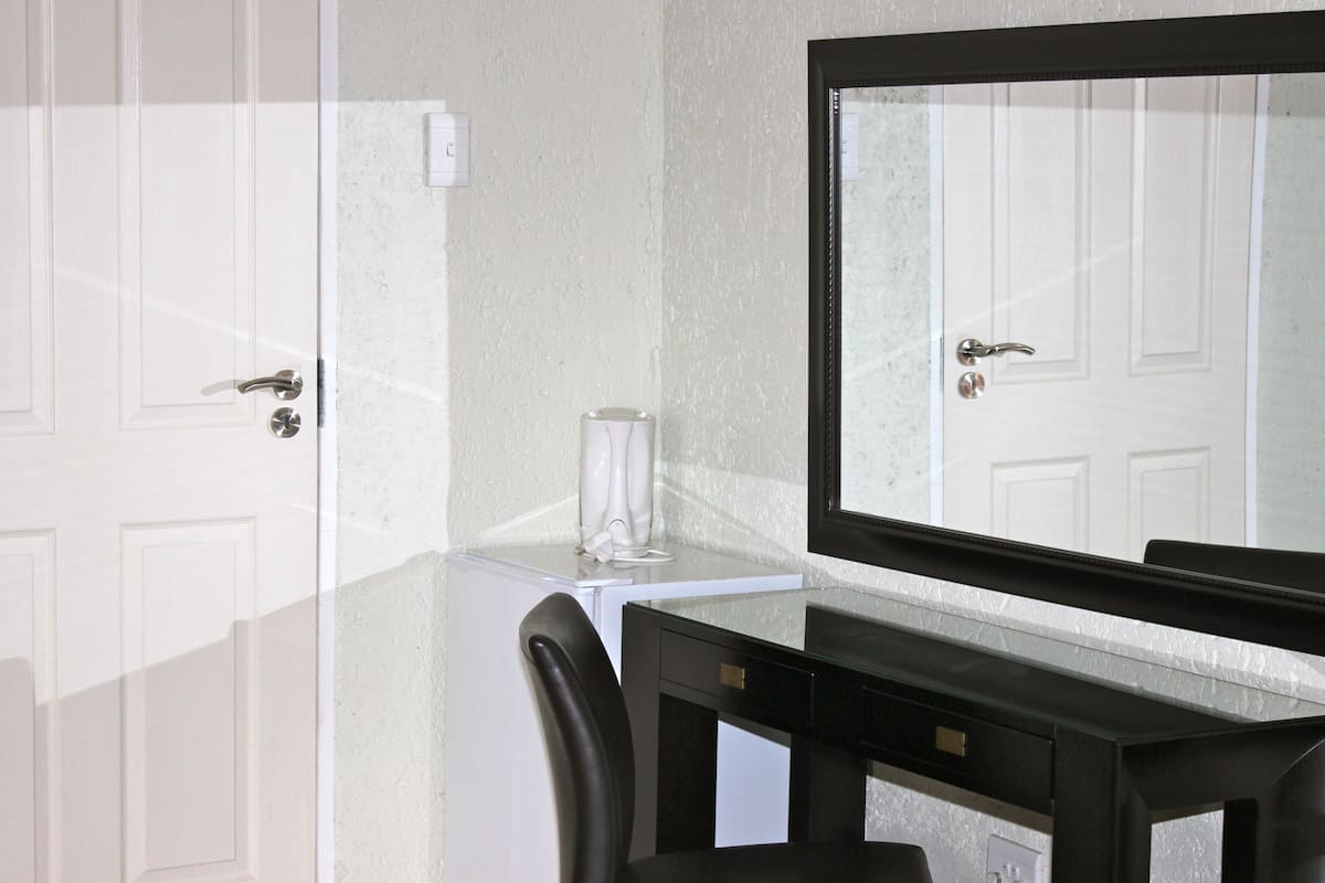 A hotel room with a white door, a small white table with a glass container, a black desk, and a large mirror.