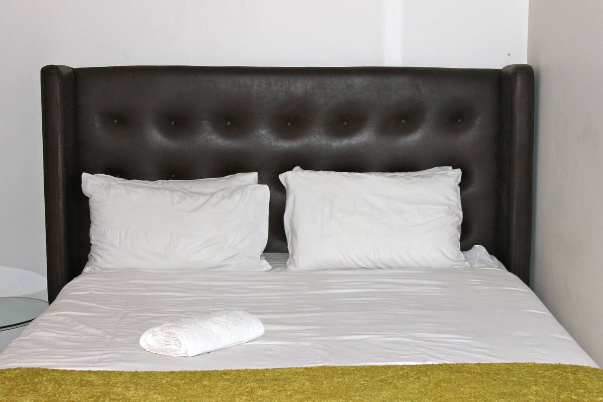 A bed with a tufted headboard, two pillows, and a rolled towel on a white bedspread.