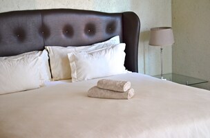 A neatly made bed with a dark headboard, white pillows, and two rolled towels.
