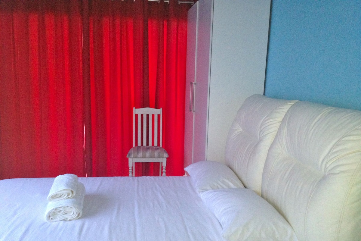 A neatly made bed with white linens and a rolled towel, a white chair, and a red curtain in the background.
