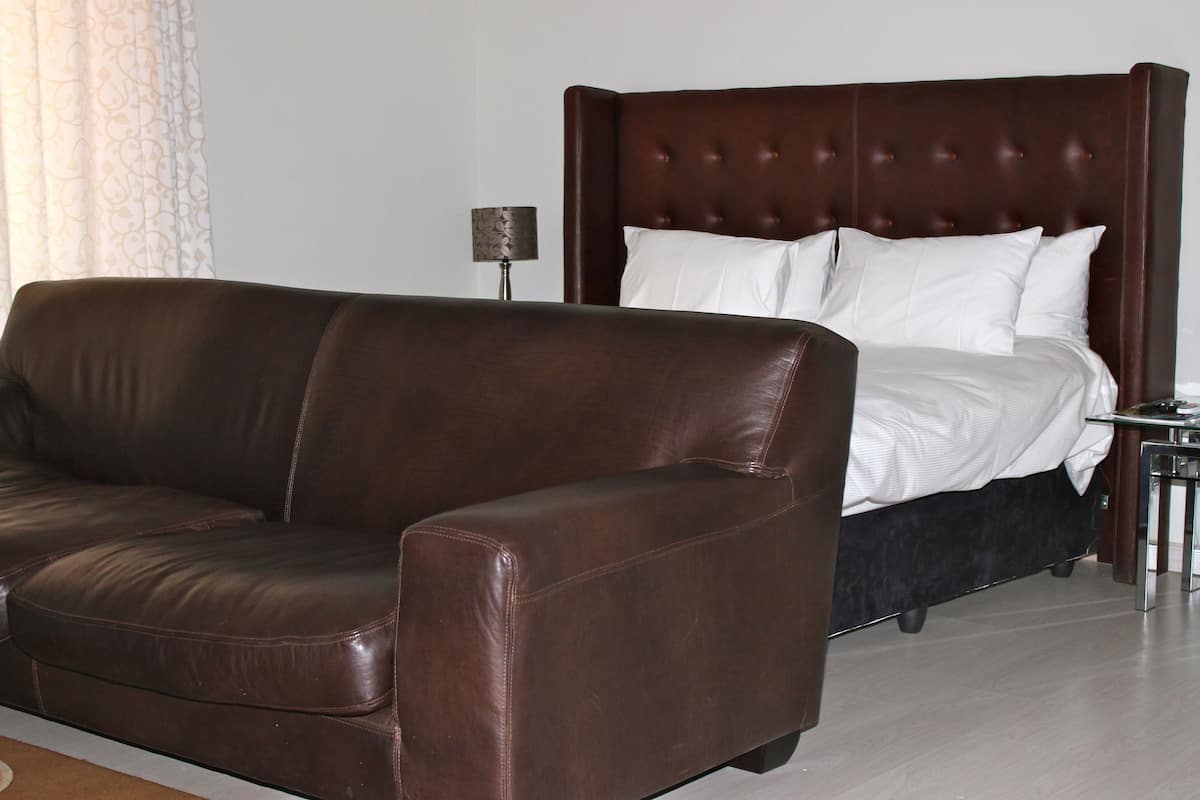 A room with a brown leather sofa, a bed with a dark headboard, and a small table with a lamp.