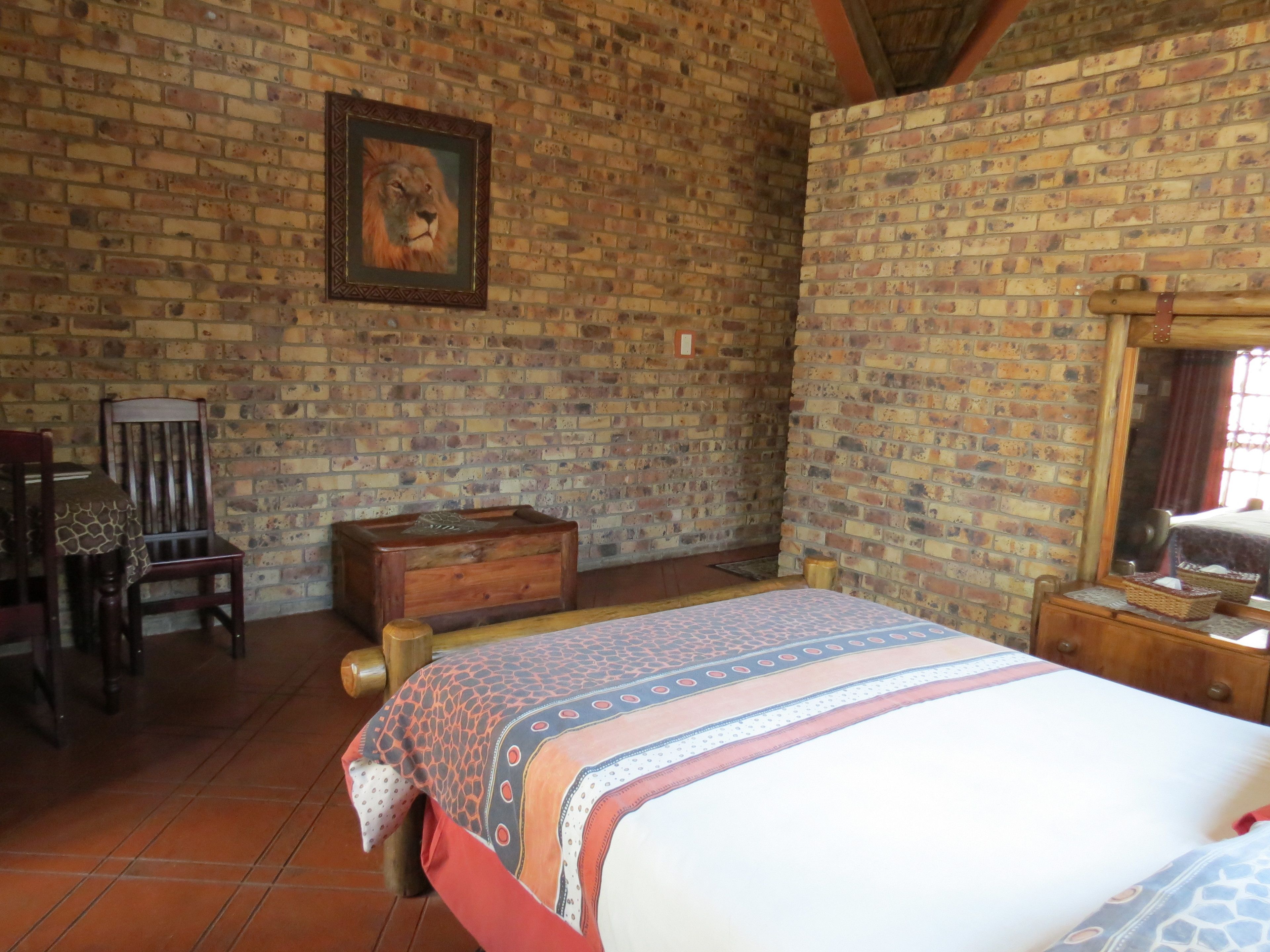 Una habitación con pared de ladrillo, cama con ropa de cama estampada, una silla de madera, un cofre de madera y un cuadro enmarcado.