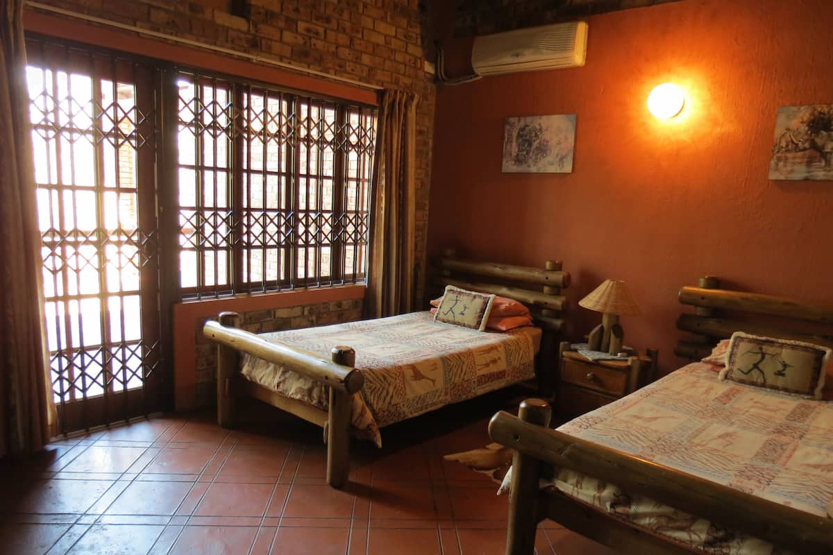 Ein Zimmer mit zwei Holzbetten, einem Fliesenboden, einem Fenster mit Metallgitter und einer Wand-Klimaanlage, sowie einem kleinen Tisch mit einer Lampe.