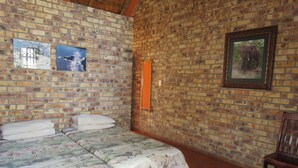 Een kamer met een bakstenen muur, een bed met een bloemenprint, een lijst met een afbeelding, en een houten stoel.
