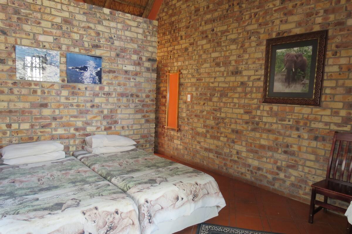 Ein Zimmer mit Backsteinwand, einem Bett mit floralem Überwurf, einem gerahmten Bild und einem Holzstuhl.