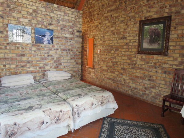 A room with a brick wall, a bed with a floral bedspread, a framed picture, and a wooden chair.