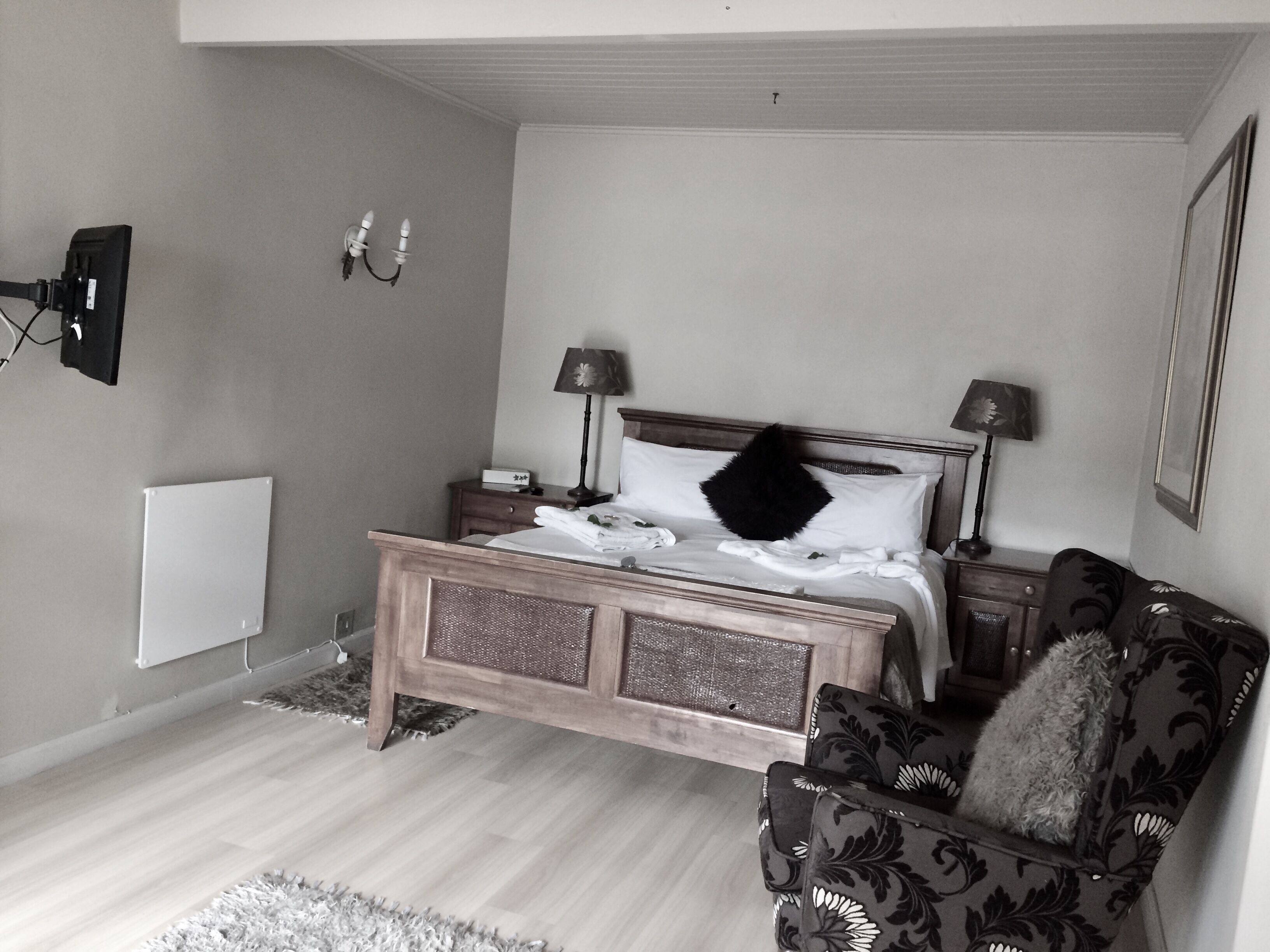 A bedroom with a wooden bed, two bedside tables with lamps, a chair with a patterned cushion, and a wall-mounted TV.