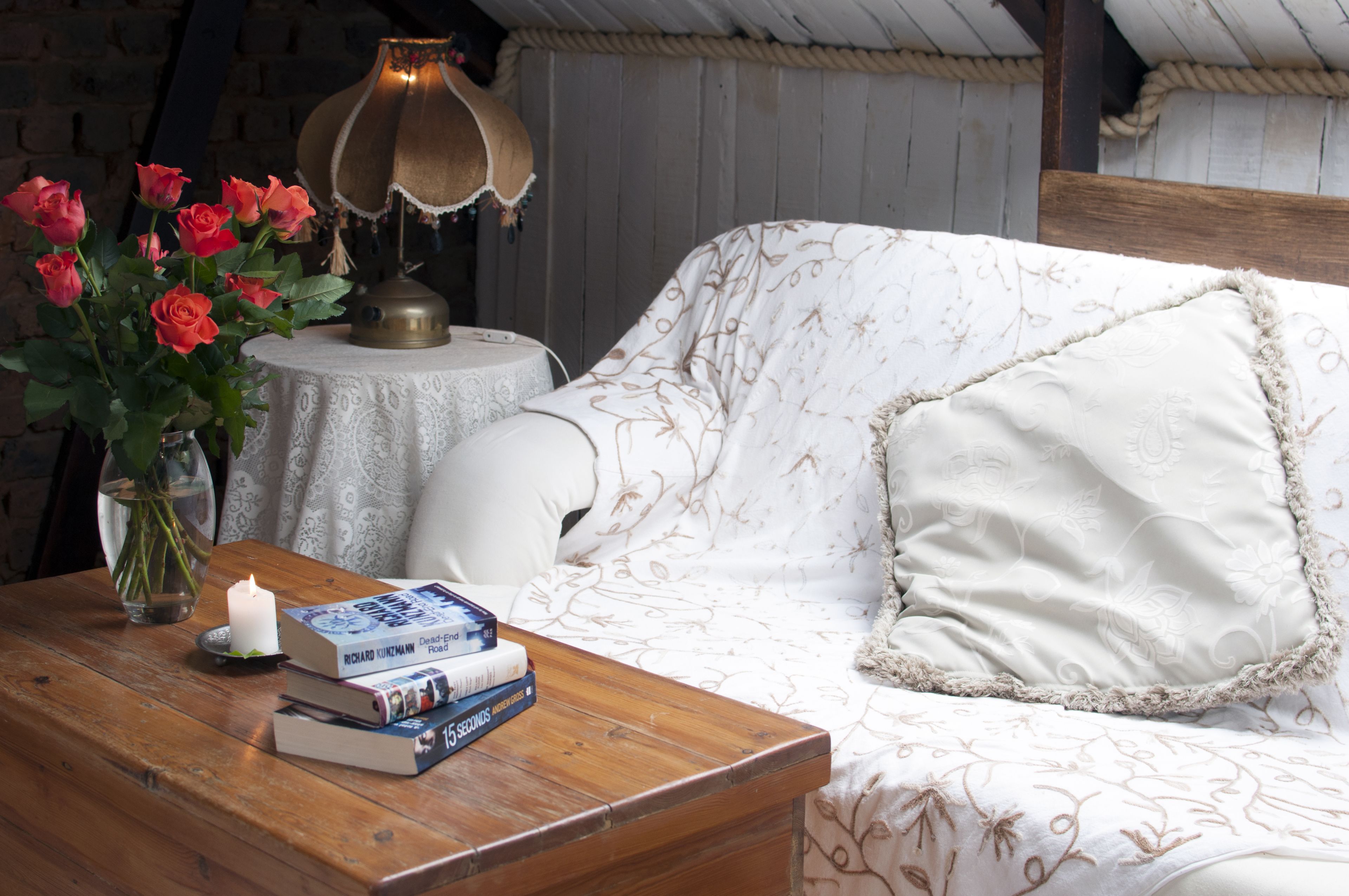 A cozy room with a wooden table, a white sofa with floral patterns, a small round table with a lamp, and a vase of red roses.