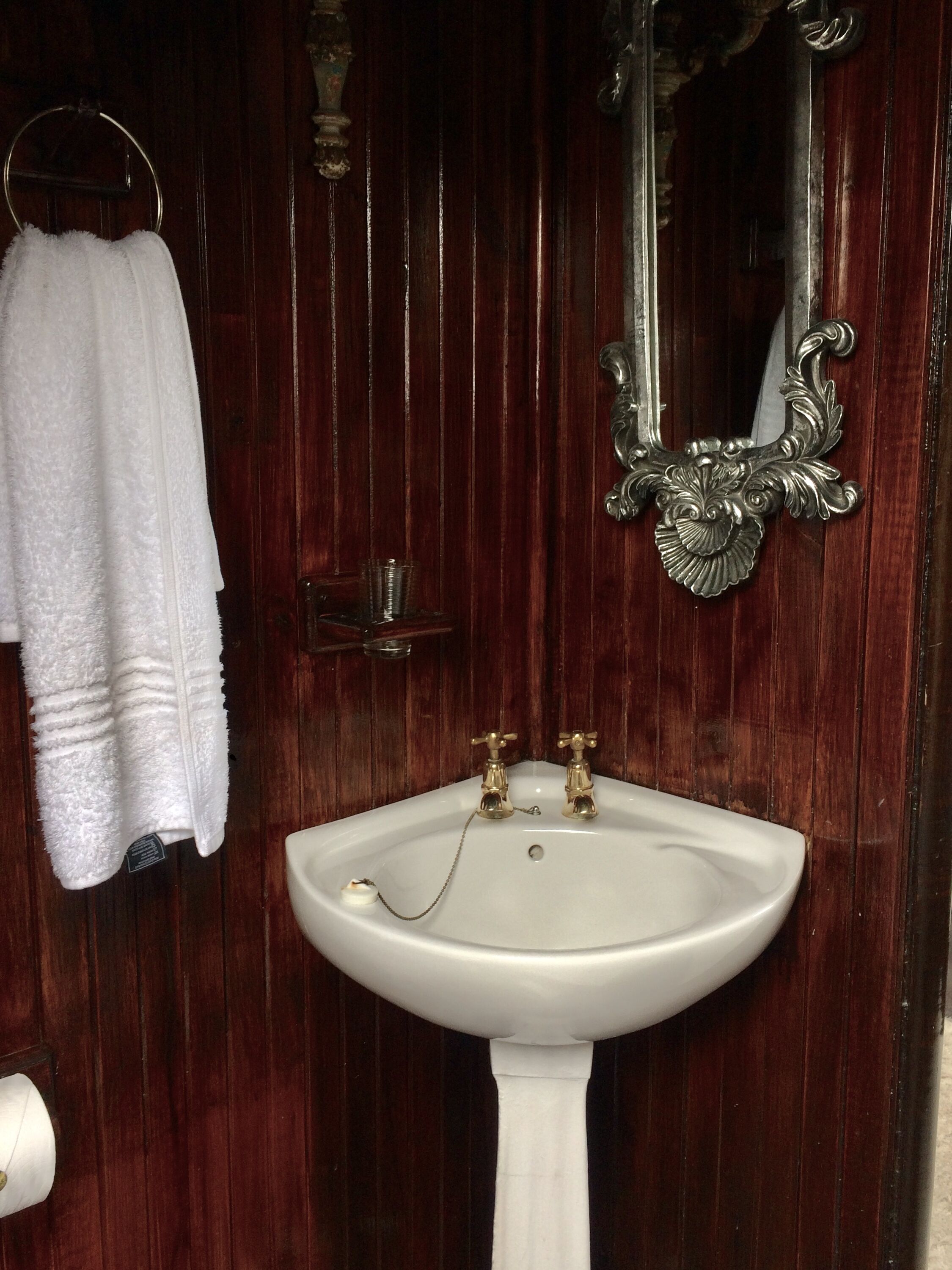 Un baño con un lavamanos blanco de pedestal, dos grifos dorados, una toalla blanca colgada en un aro de metal y un espejo con un marco ornamentado.