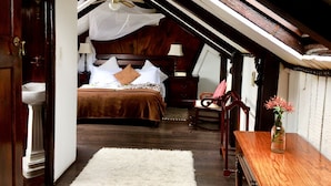 A bedroom with a wooden ceiling, a bed with white linens, a wooden chair, a wooden table with a vase, and a white rug.