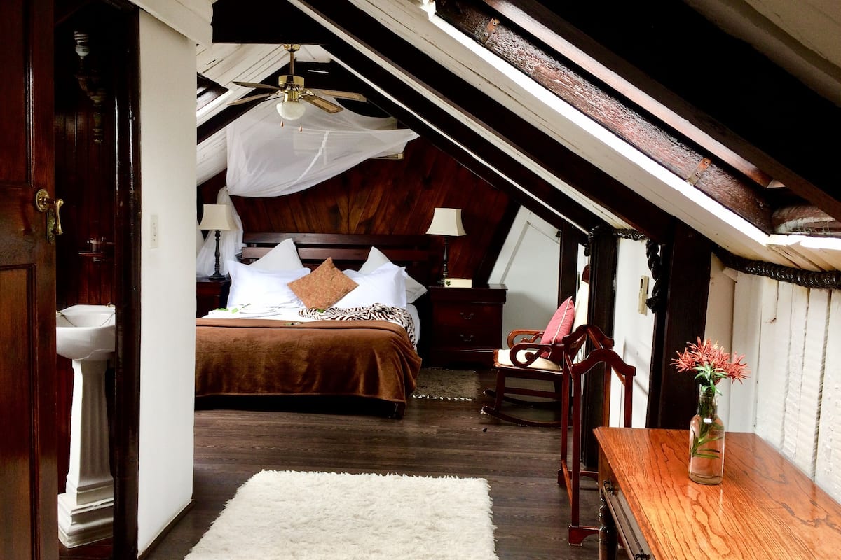 Quarto com teto de madeira, cama com roupa de cama branca, cadeira de madeira, mesa de madeira com um vaso e tapete branco.