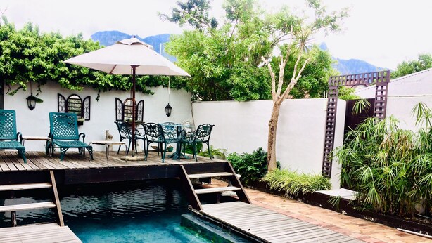 A pool area with wooden decks, a table and chairs, and a white wall with greenery.