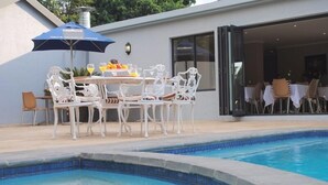 Un espace repas aménagé au bord de la piscine, avec des chaises blanches et une table, un parasol et un plateau de fruits.