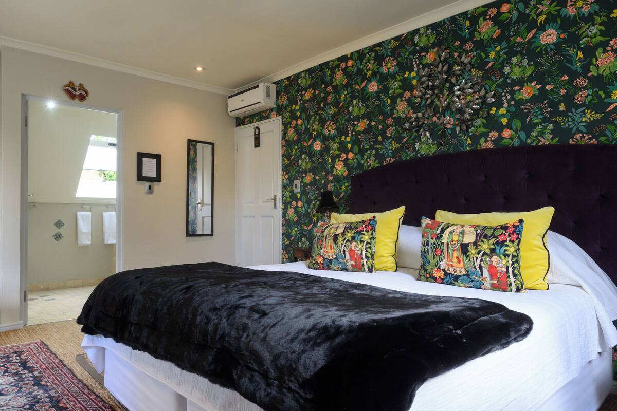 A bedroom with a large bed, floral wallpaper, a dark headboard, and a visible air conditioning unit.