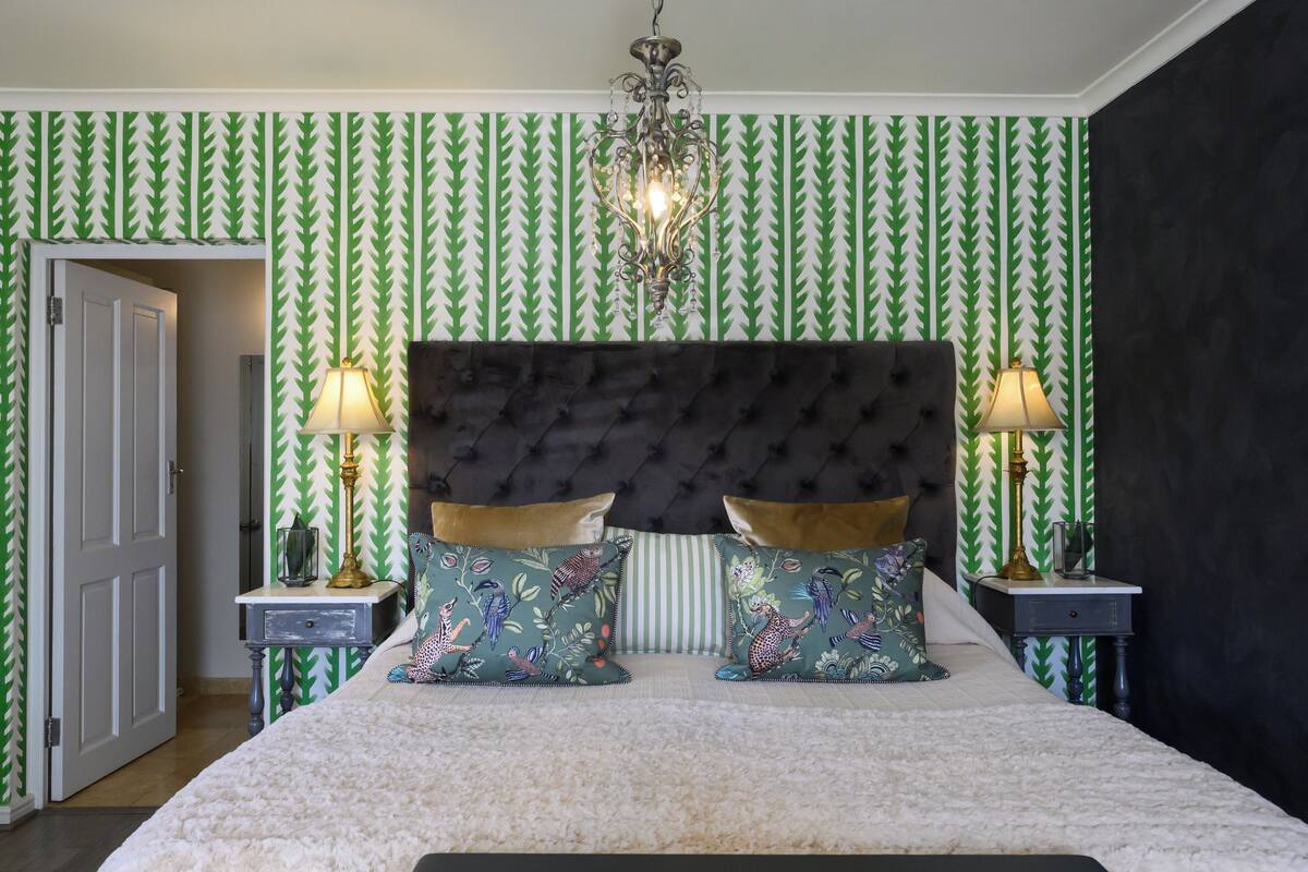 A bedroom with a tufted headboard, two bedside tables with lamps, patterned pillows, and a chandelier.
