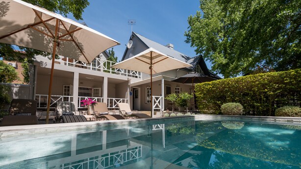 A swimming pool with lounge chairs and umbrellas, a covered patio area, and a building with a sloped roof. - Baruch Guest House on Rhodes Noord (Stellenbosch)
