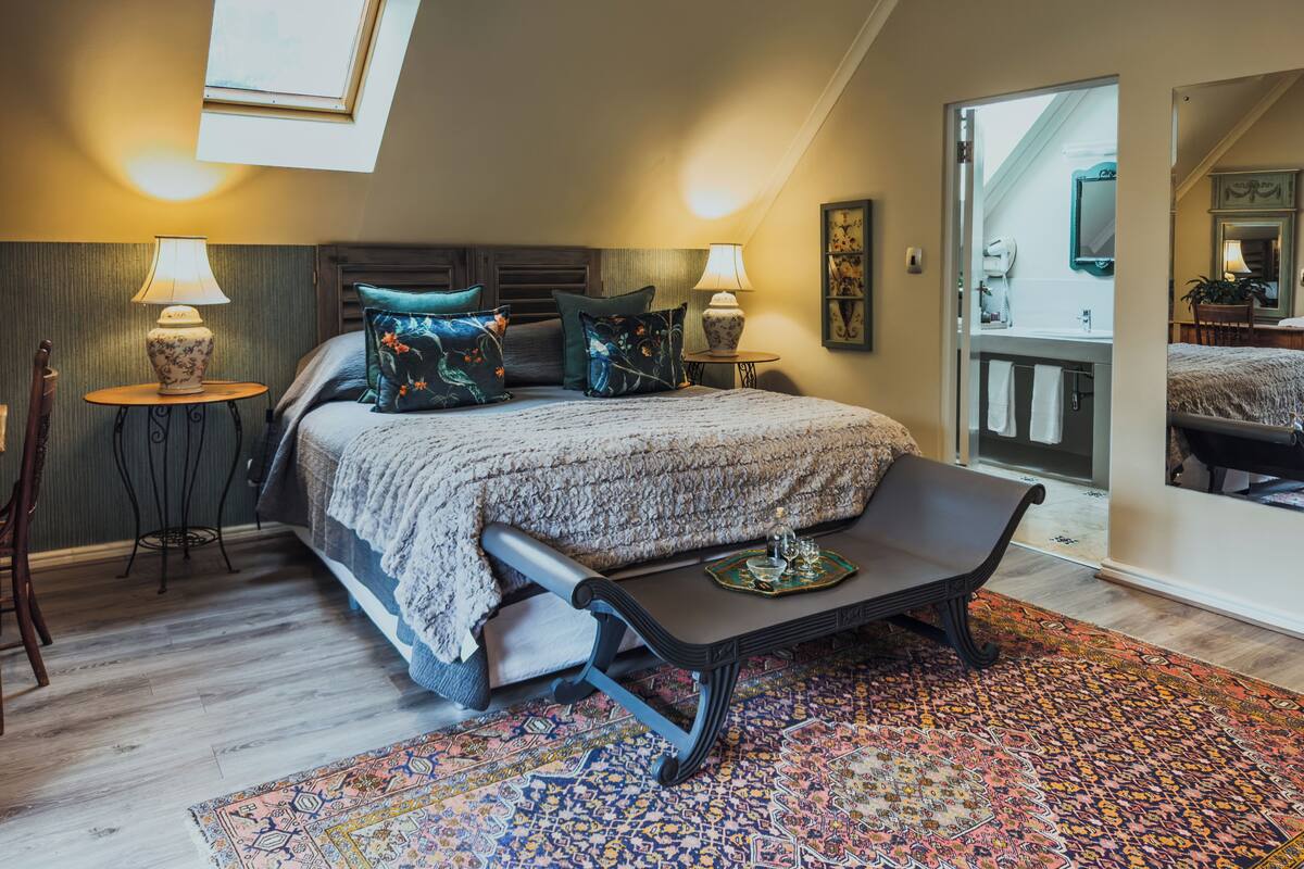 A bedroom with a bed, two bedside tables with lamps, a bench, a patterned rug, and a skylight.