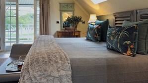 A bedroom with a large bed, a desk with a tray of glasses and a bottle, a lamp, and a view of the outside through a window. - Baruch Guest House on Rhodes Noord (Stellenbosch)