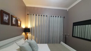 A hotel room with a bed, a nightstand with a lamp, a window with curtains, and framed pictures on the wall. - 2 Leafed Doors (Sandton)
