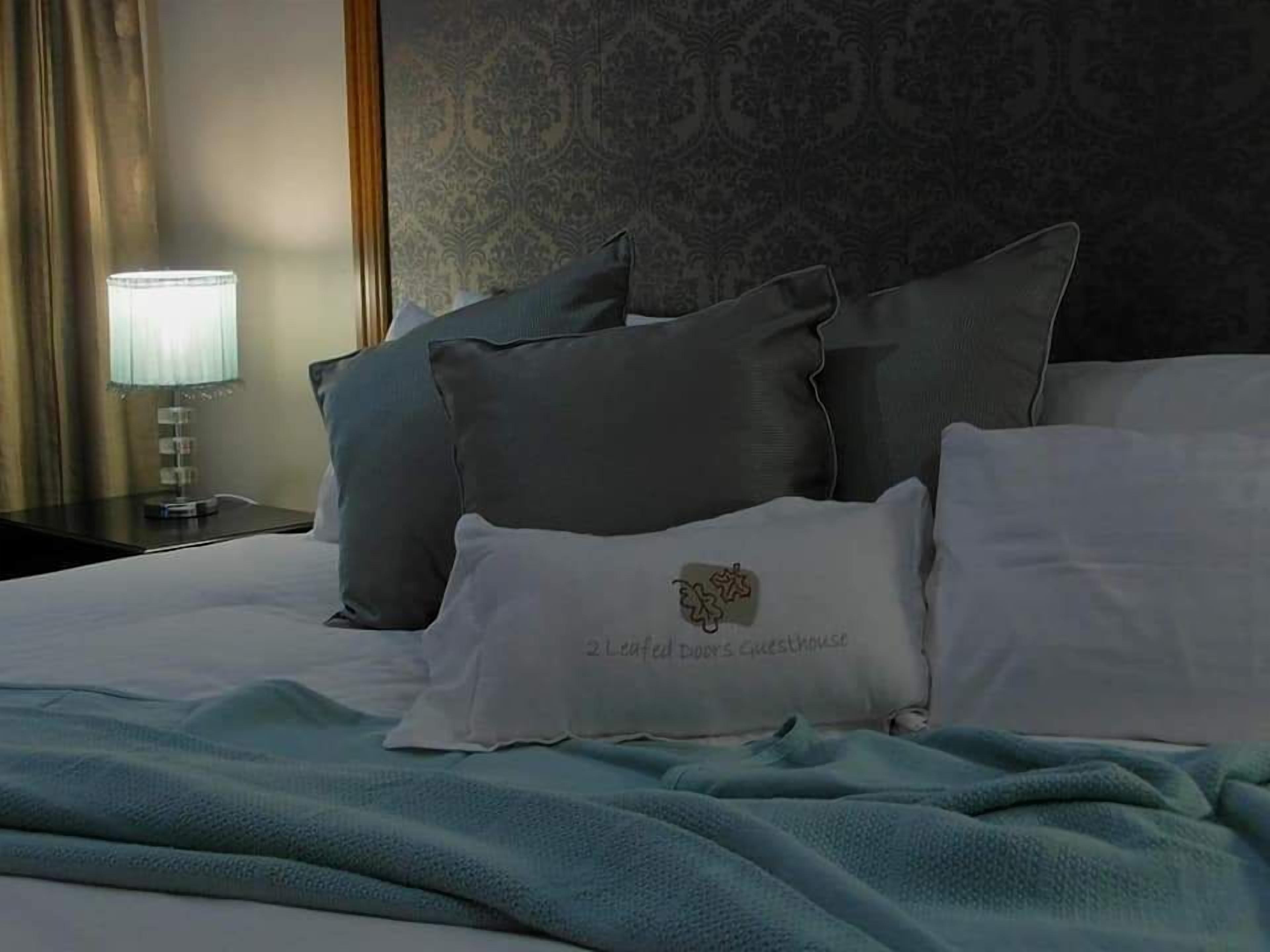 Un lit bien fait, avec des oreillers gris et blancs, une couverture bleue et une table de chevet sur laquelle se trouve une lampe.