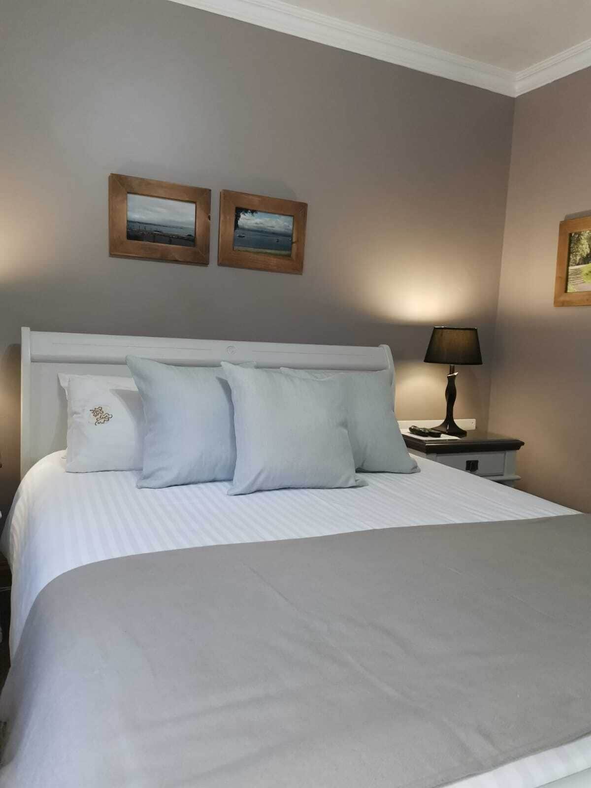 Un lit bien fait, avec du linge de lit blanc et plusieurs oreillers, une lampe de chevet et deux tableaux encadrés accrochés au mur.