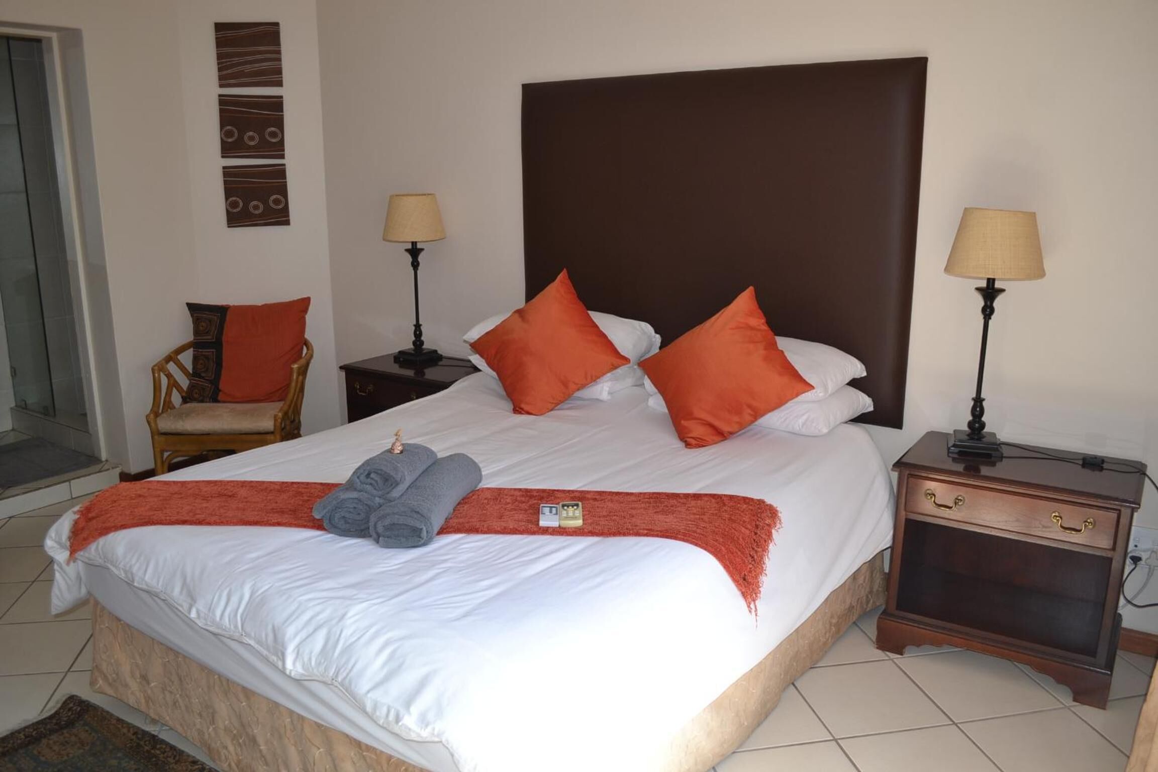 A neatly made bed with white linens, orange pillows, and a red throw blanket. There are two bedside tables with lamps, a wooden chair, and a small rug on the floor.