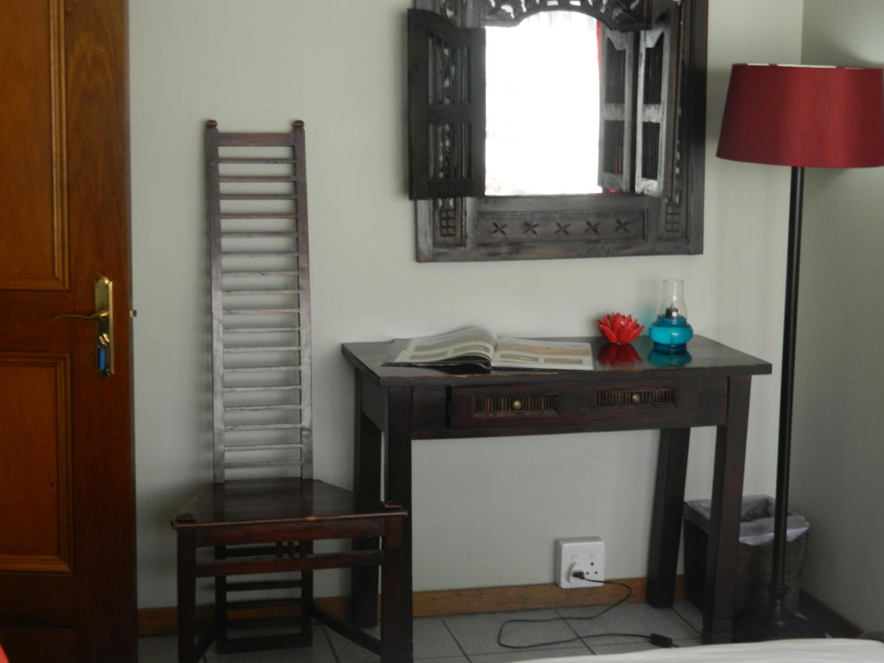 Une pièce avec une chaise en bois, un bureau sur lequel se trouve un livre, une lampe et un miroir accroché au mur.