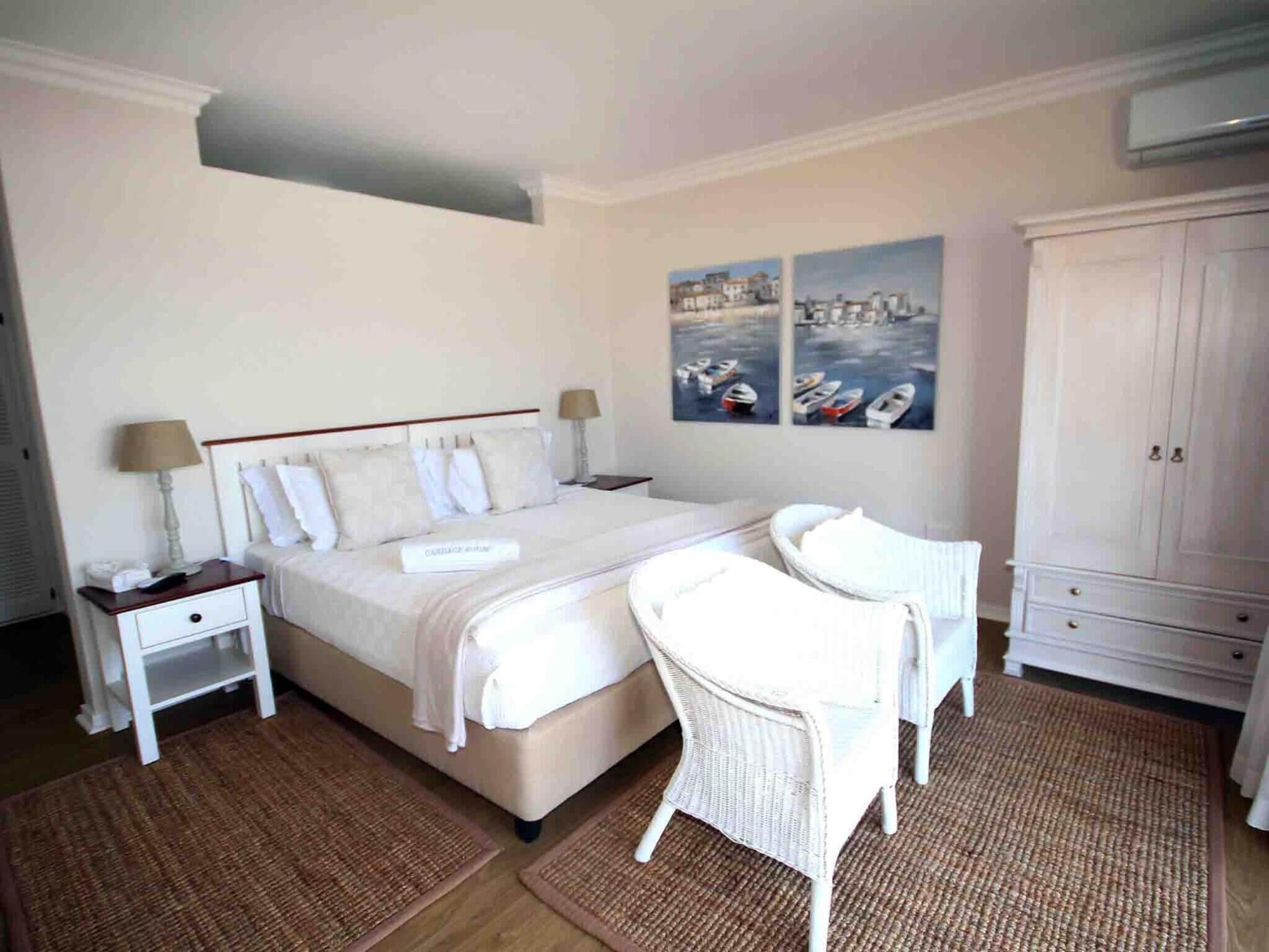 Un dormitorio con cama, mesitas de noche, una silla, un armario y dos cuadros en la pared.