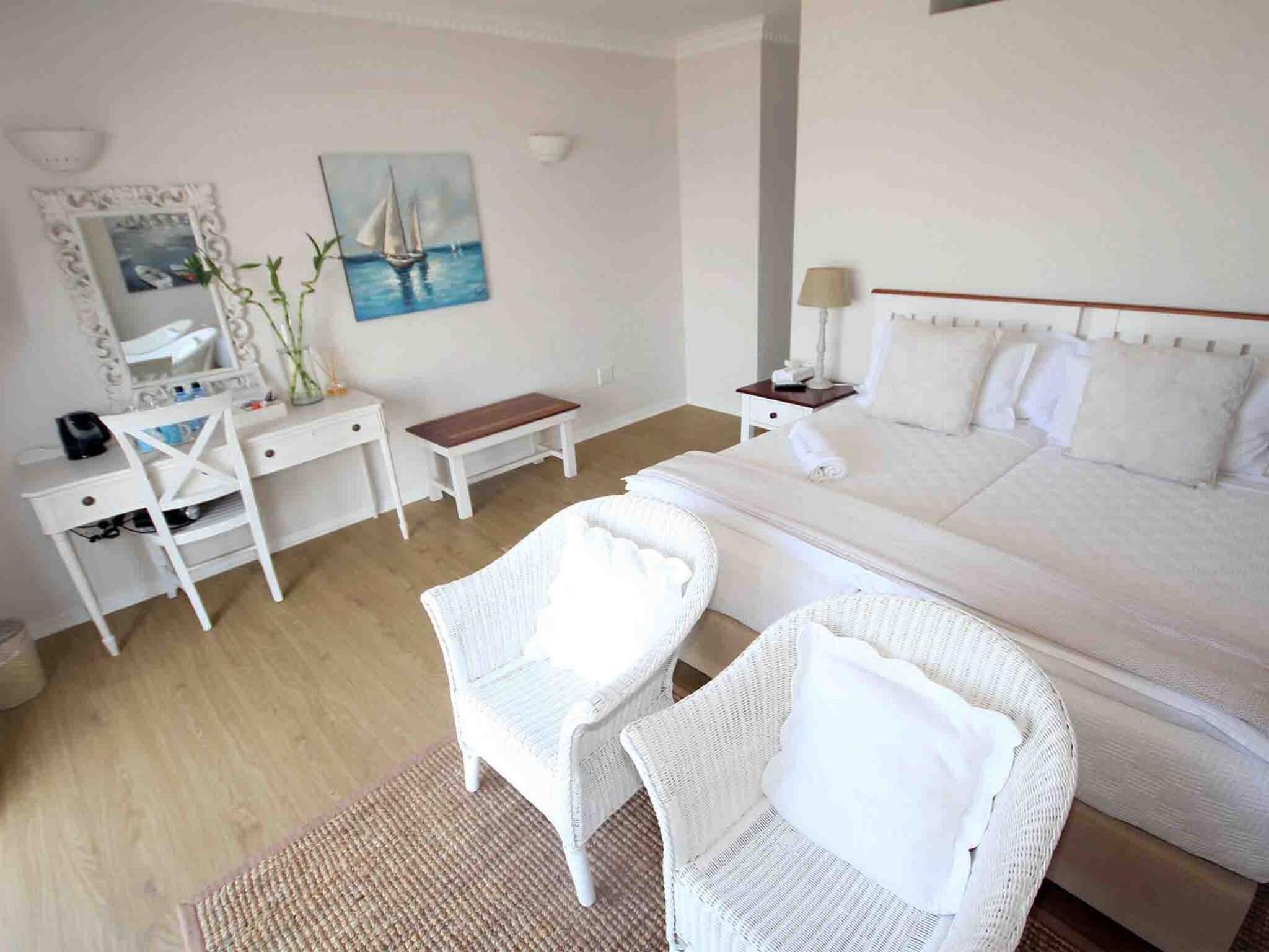 Habitación de hotel con cama, dos sillas de mimbre blancas, un escritorio con espejo, una mesita y un cuadro de un velero.