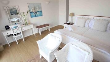 Habitación de hotel con cama, dos sillas de mimbre blancas, un escritorio con espejo, una mesita y un cuadro de un velero.